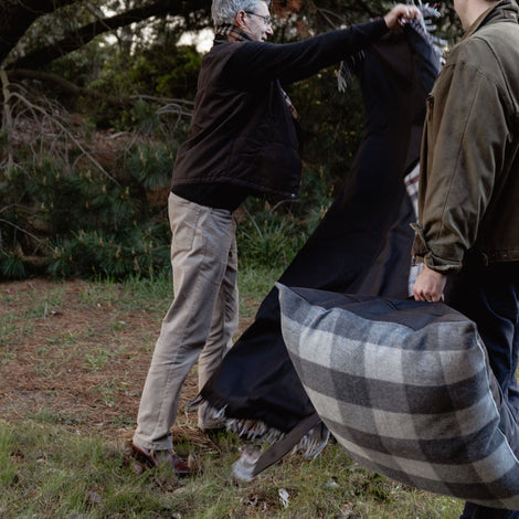 Two people outdoors, one holding a plaid blanket, with trees in the background.