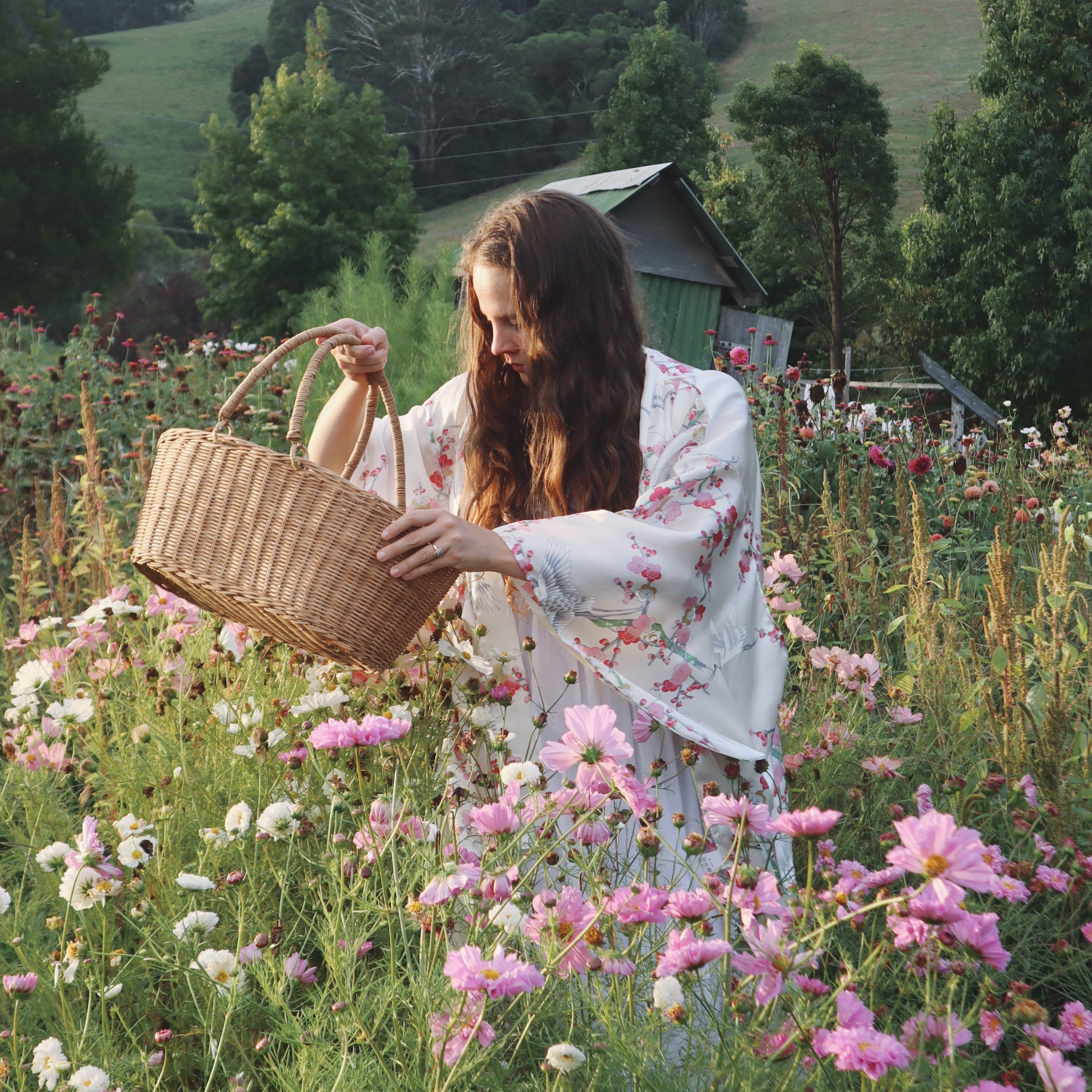 Woman in a floral dress holding a woven basket in a field of flowers with mountains in the background
