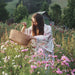 Woman in a floral dress holding a woven basket in a field of flowers with mountains in the background