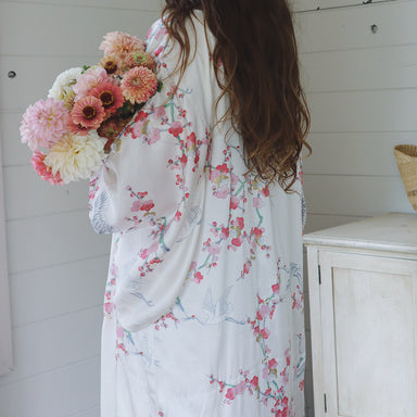 Woman in a floral kimono robe holding flowers in a room with white walls and a framed picture.