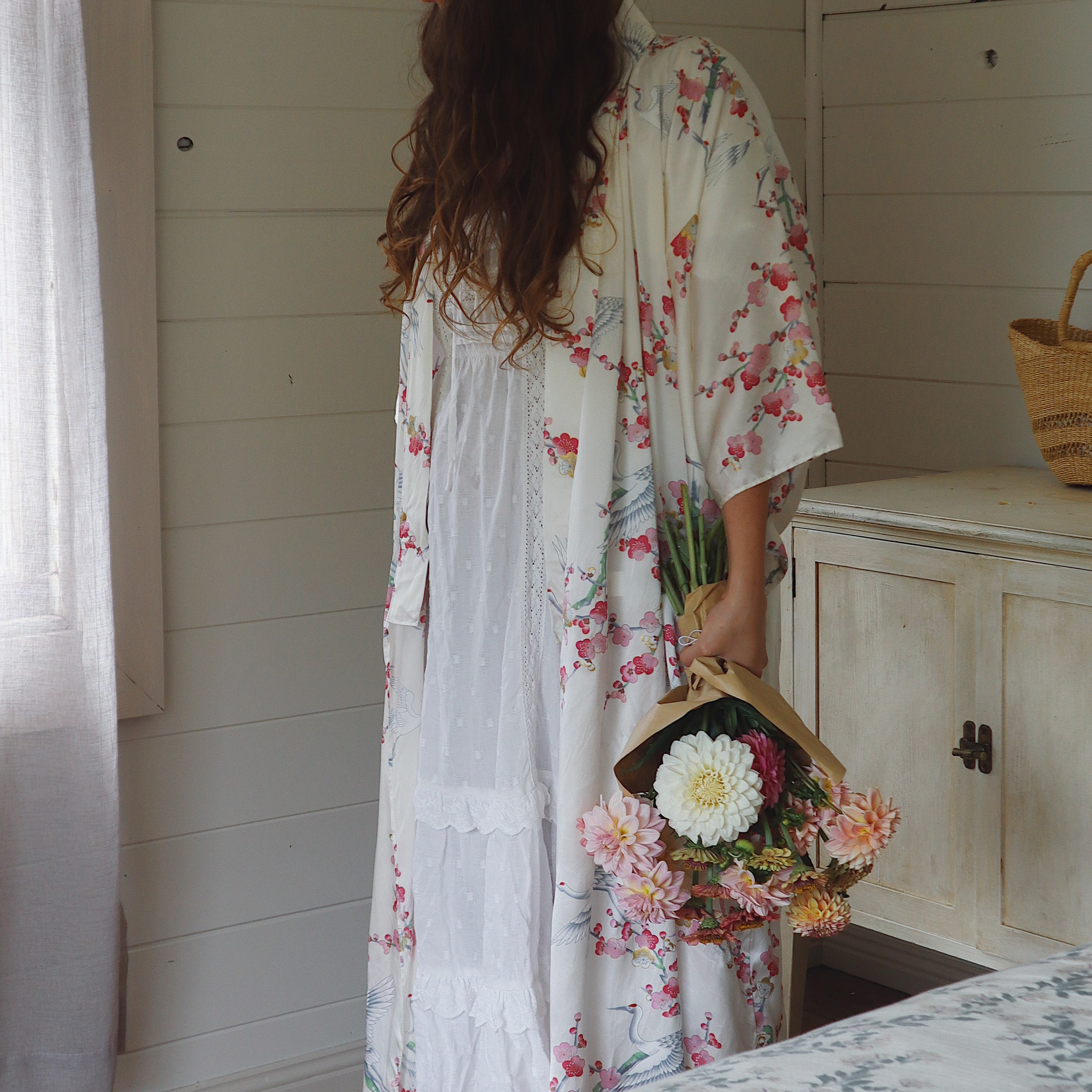 Woman in a floral kimono robe standing by a bed holding a basket of flowers in a room with white walls.