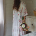 Woman in a floral kimono robe standing by a bed holding a basket of flowers in a room with white walls.