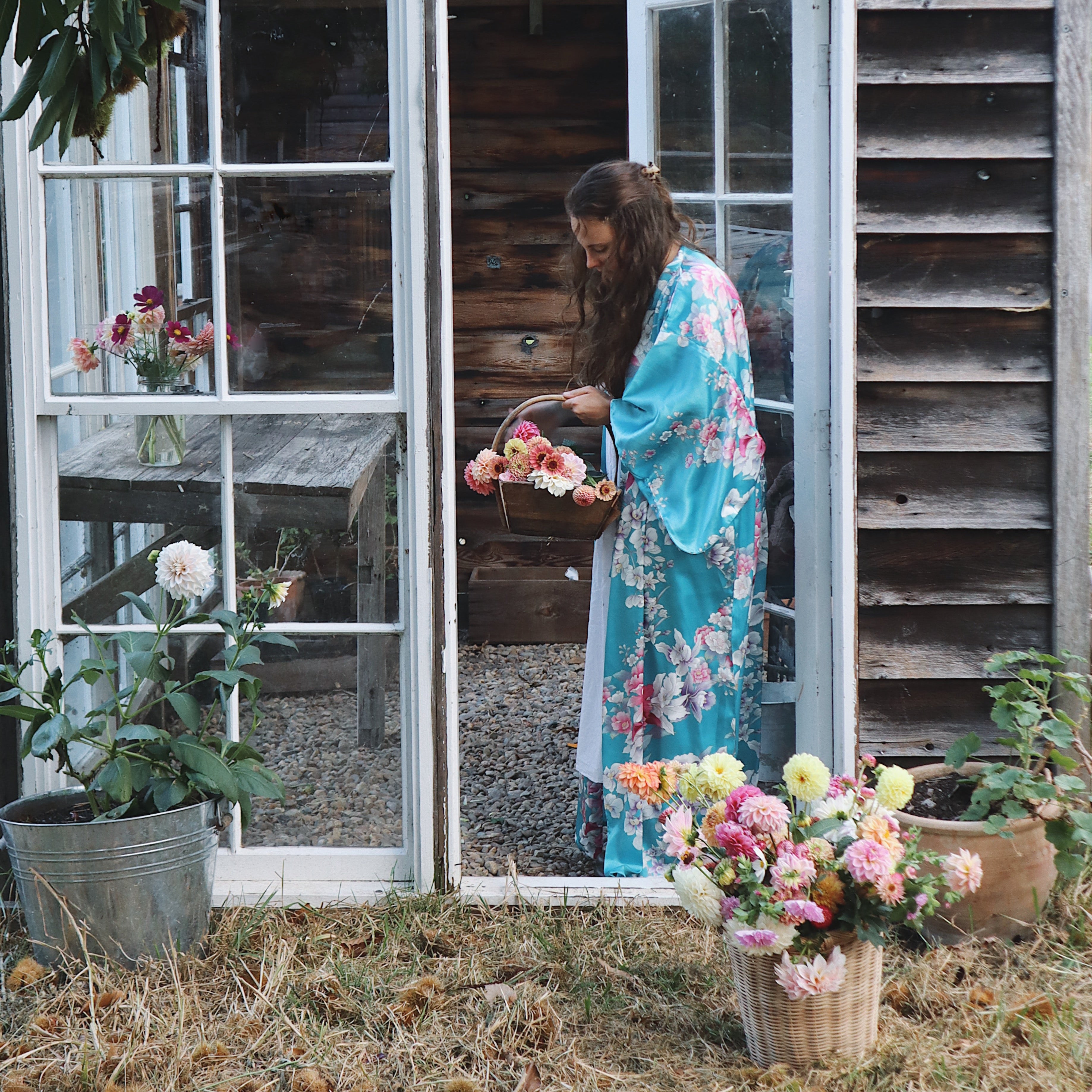 Woman in a floral kimono standing in front of a wooden shed with flowers around her.