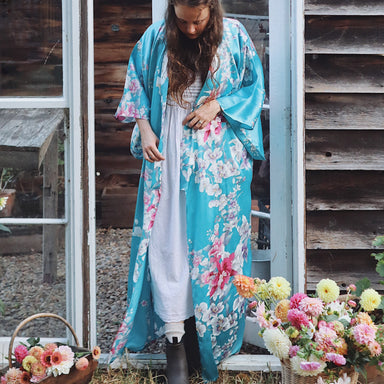 Woman in a floral kimono standing outdoors with flowers and a wooden shed in the background