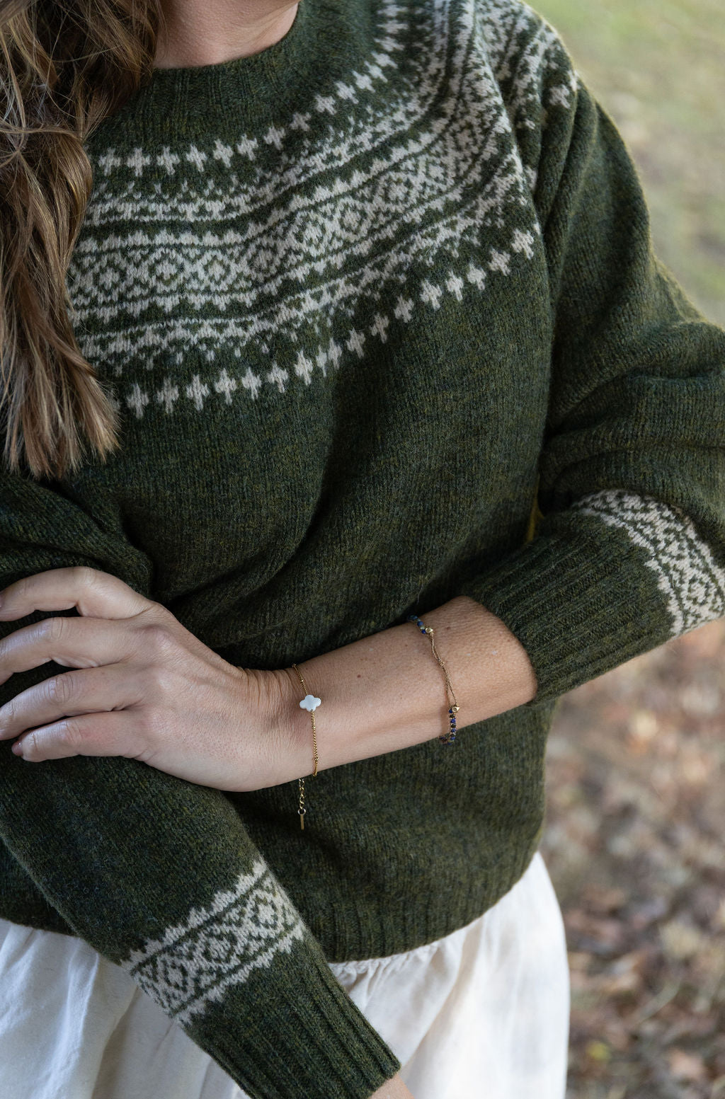 Person wearing a green fair isle knit sweater with a pattern, holding their arm with a bracelet.