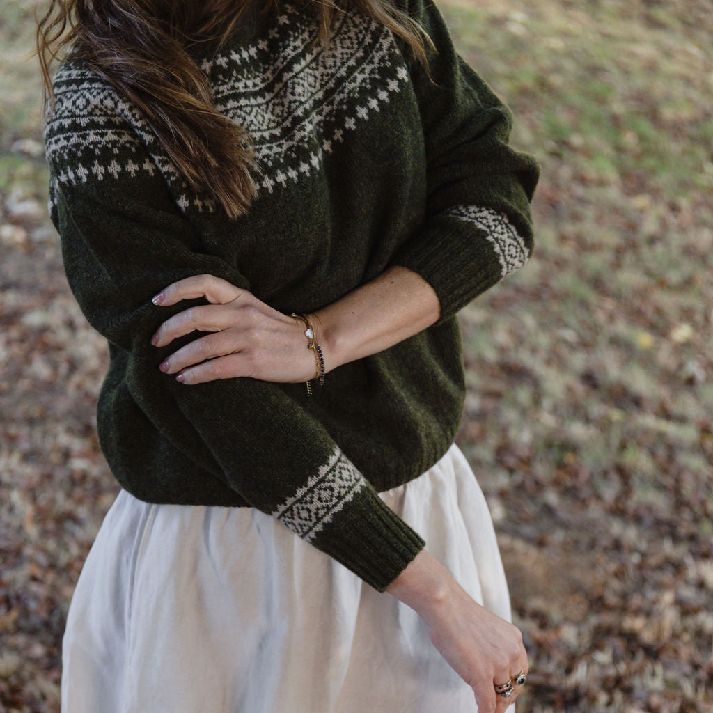 Person wearing a dark green fair isle knit sweater with a white pattern and a white skirt outdoors.