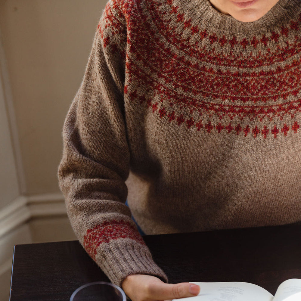 Person wearing a patterned sweater with a glass of red wine and an open book on a table.