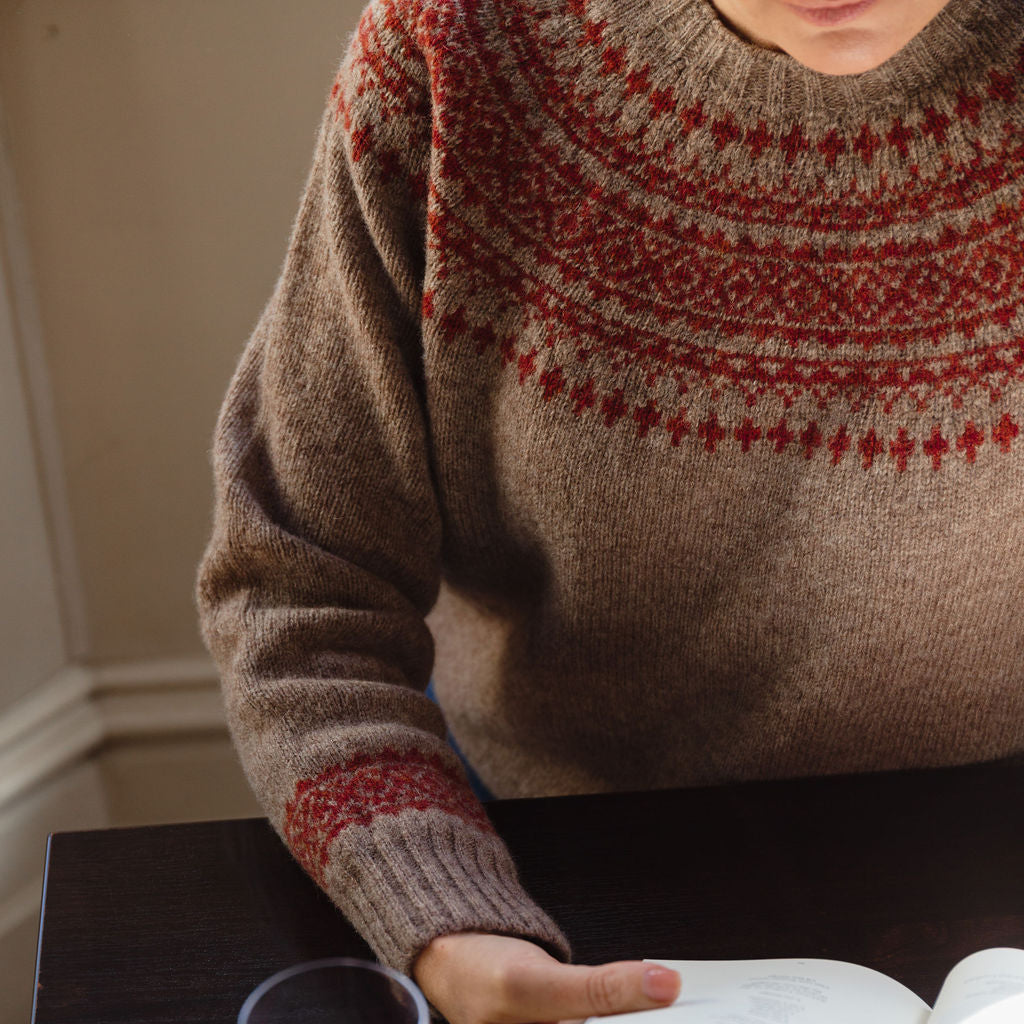 Person wearing a patterned sweater with a glass of red wine and an open book on a table.