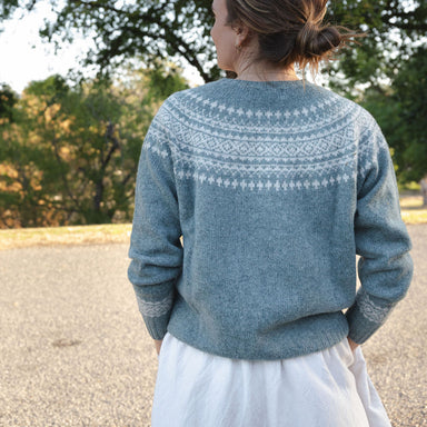 Person wearing a blue sweater with a pattern, standing outdoors with trees in the background