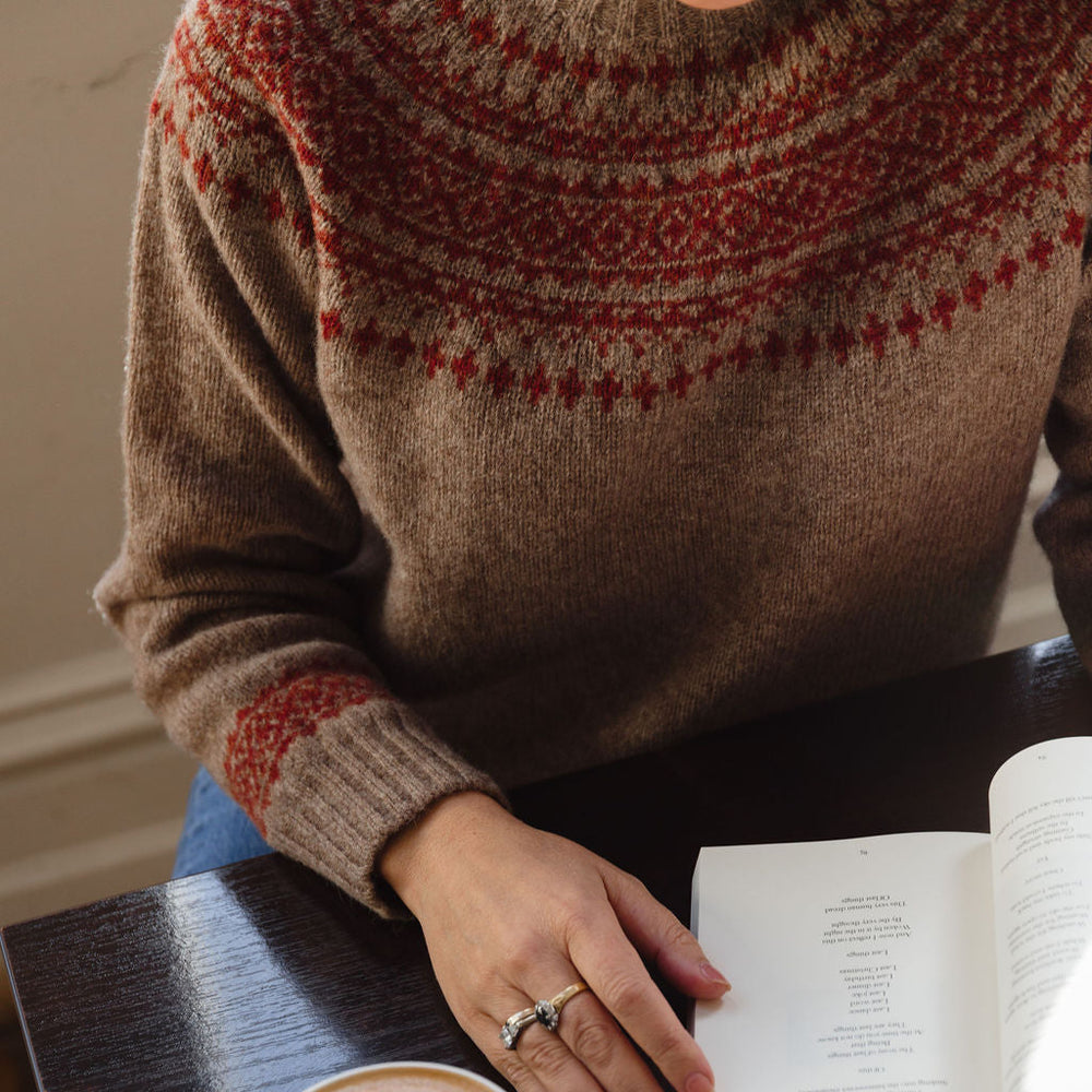 Person wearing a patterned sweater with a cup of coffee and an open book on a table.