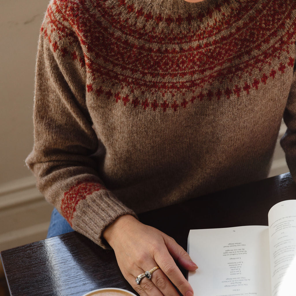 Person wearing a patterned sweater with a cup of coffee and an open book on a table.