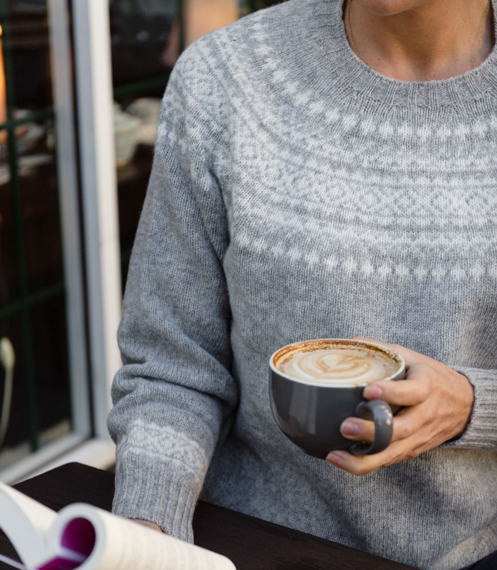 Person in a fair isle knit sweater holding a cup of coffee and a magazine outdoors