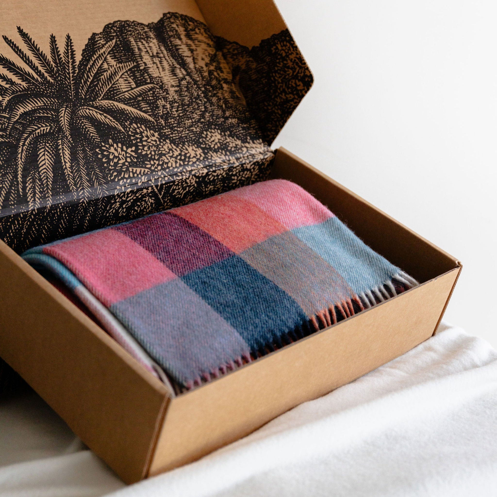Open cardboard box containing a patterned wool blanket and a striped fabric on a white surface.