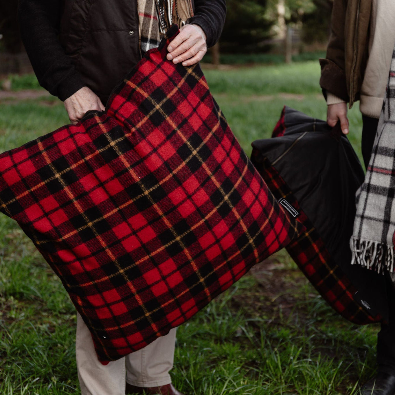 Two people holding plaid blankets outdoors on a grassy area.