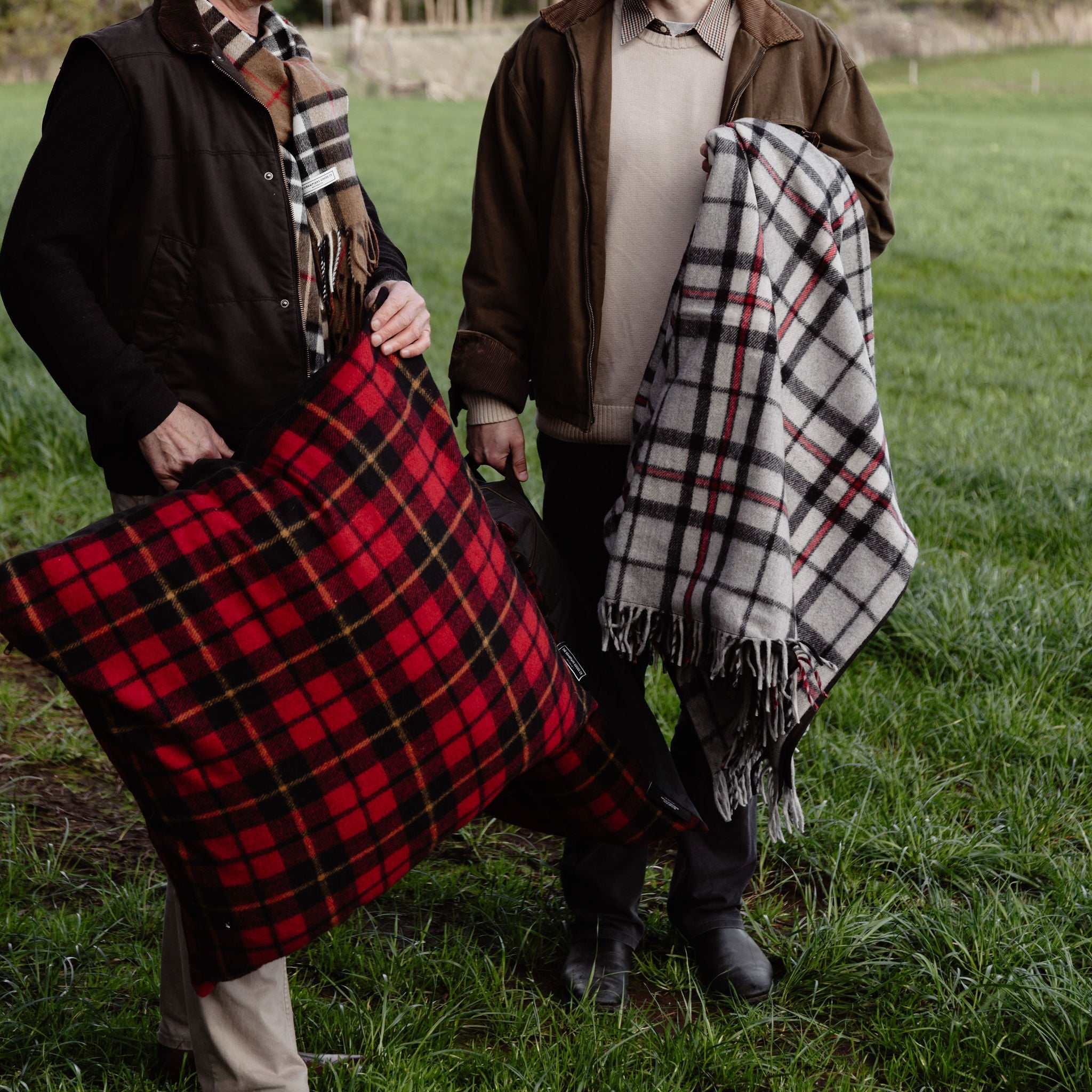 Two men standing in a grassy field, one holding a red tartan pillow and the other with a plaid blanket.