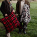 Two men standing in a grassy field, one holding a red tartan pillow and the other with a plaid blanket.