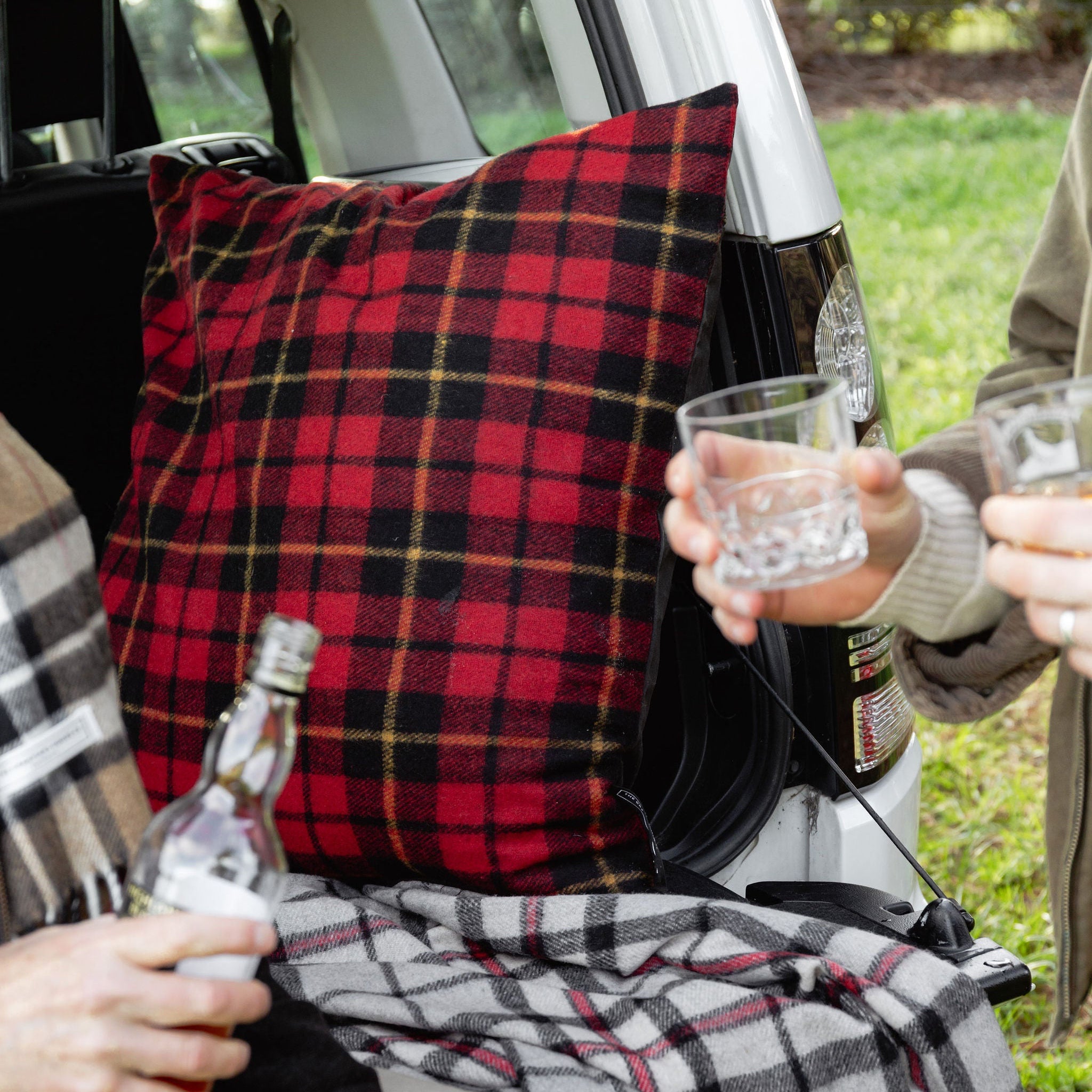 Two people sitting in a car with plaid blanket and pillow, holding drinks.
