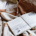 Open book with illustrations on a patterned blanket with a teddy bear and wooden spoon.