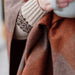 Close-up of a person wrapped in a checkered rust and brown wool blanket with a patterned sleeve and holding a white mug.