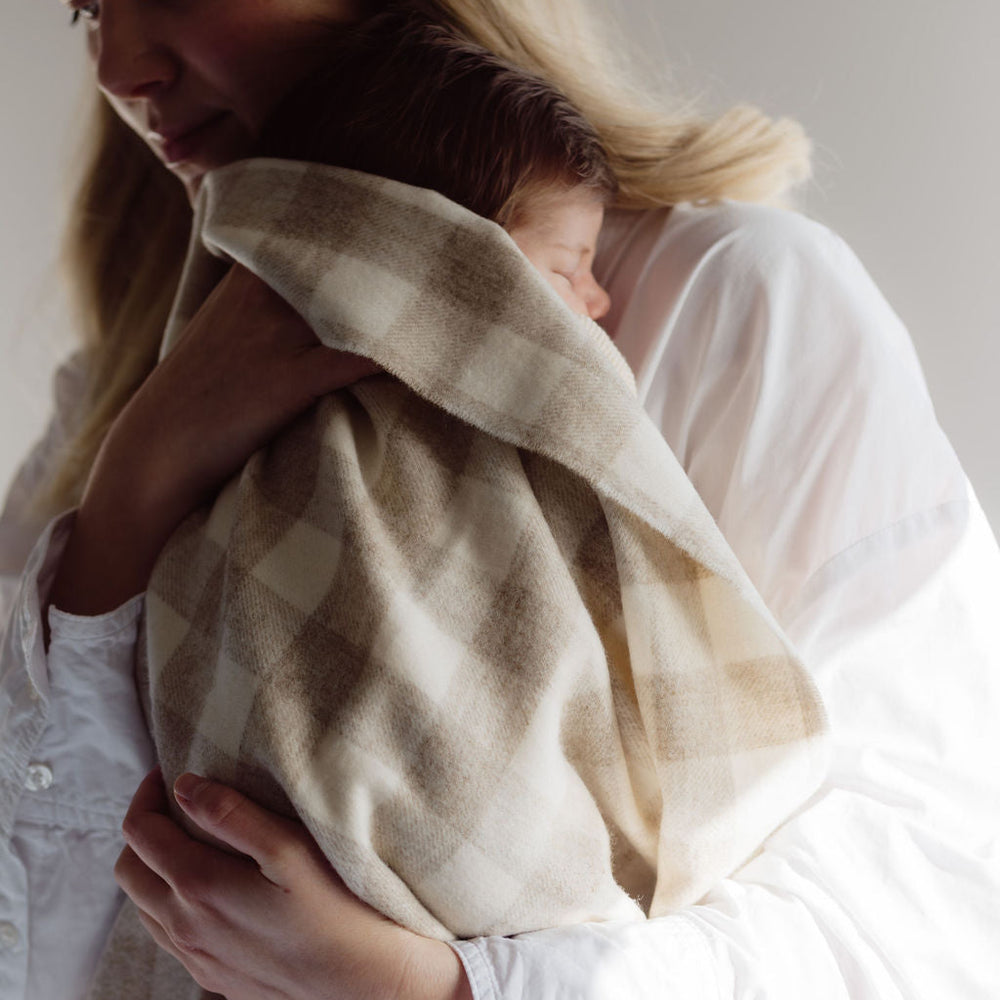 Woman holding a baby wrapped in a beige blanket against a plain background