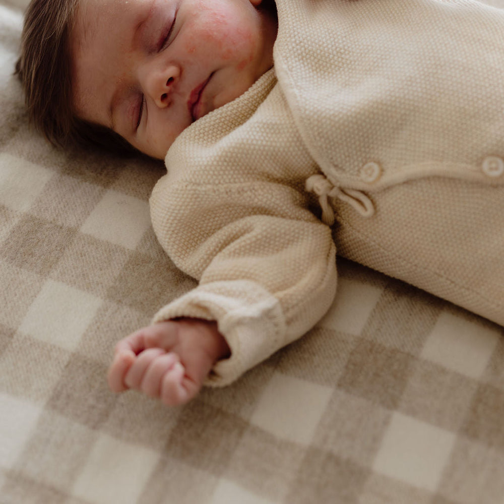 Baby in a cream-colored outfit being comforted by an adult hand on a checkered blanket.