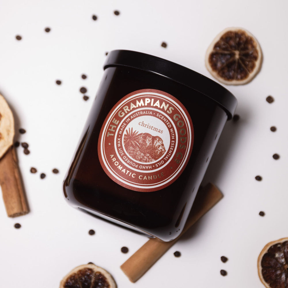 Black candle jar with a red label on a white surface with dried oranges and cinnamon sticks.