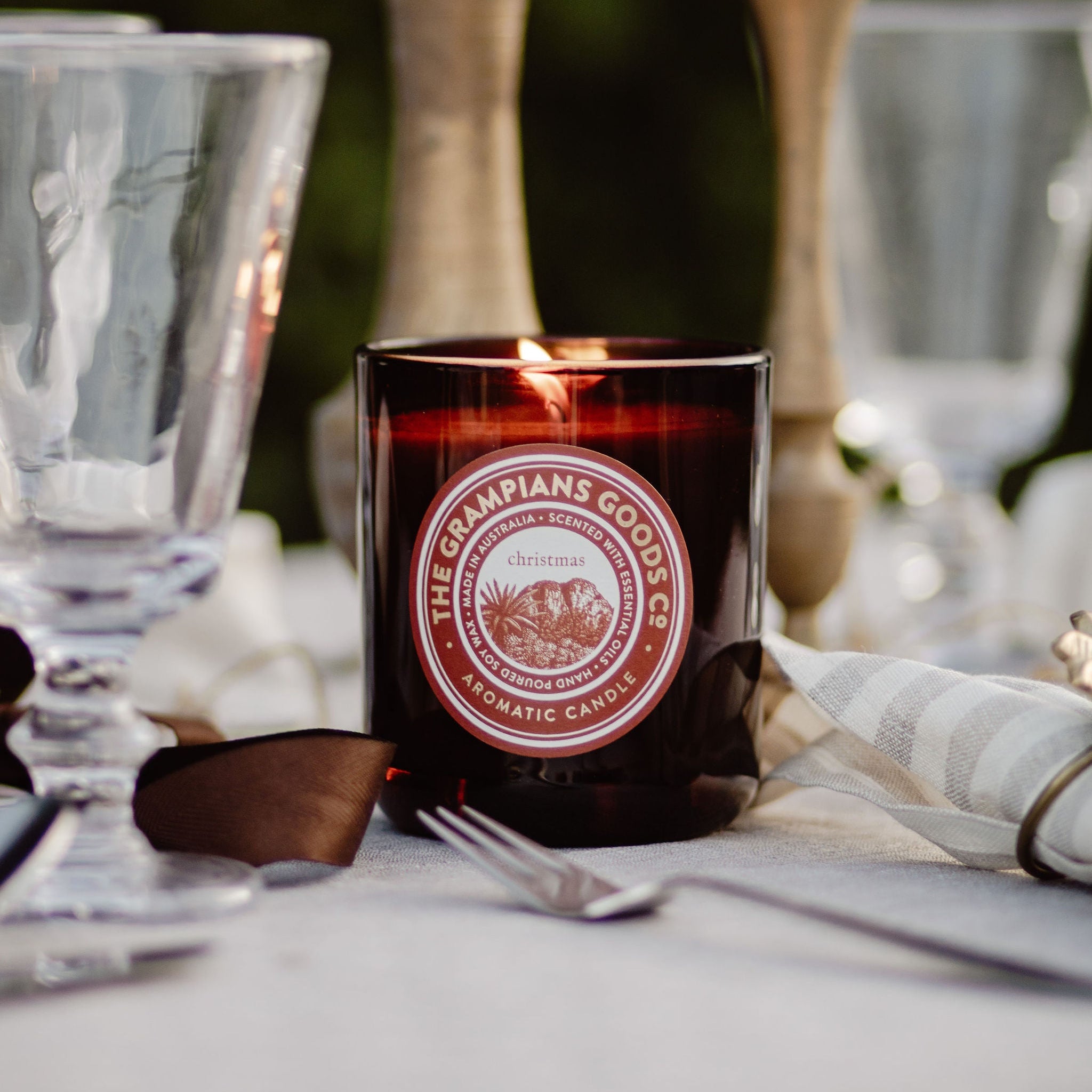 Candle with 'The Grampians Goods Co' label on a table setting with glasses and flowers.