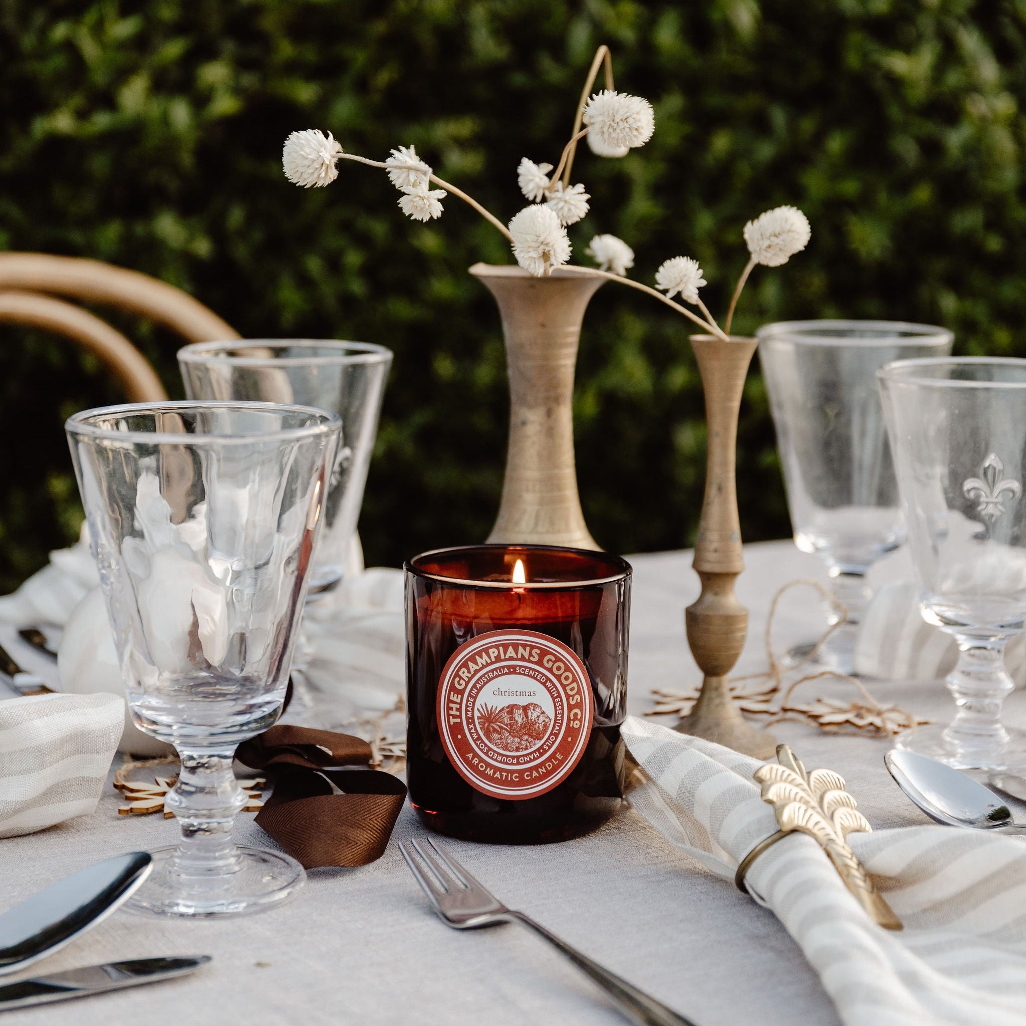 Set table with candles, glasses, and a vase with flowers against a green hedge background