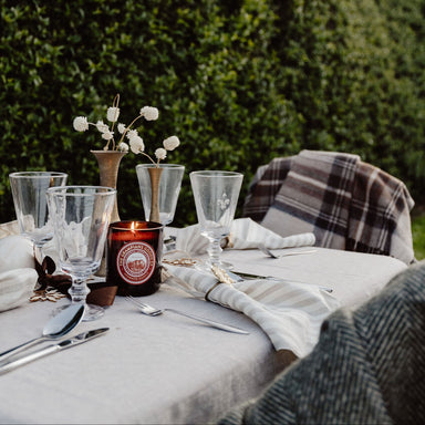Outdoor table setting with glasses, candle, and plaid blanket against a green hedge background