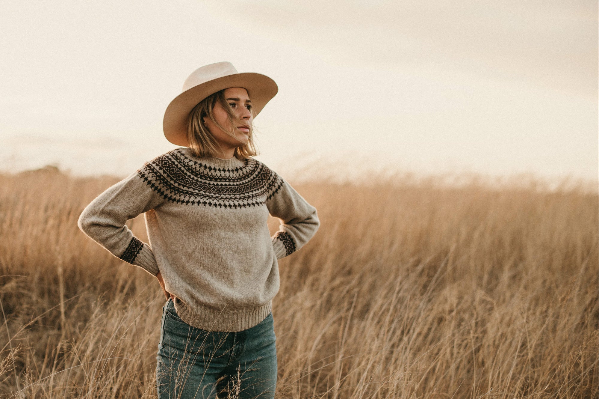 Woman wearing a patterned sweater and hat standing in a field with a neutral background