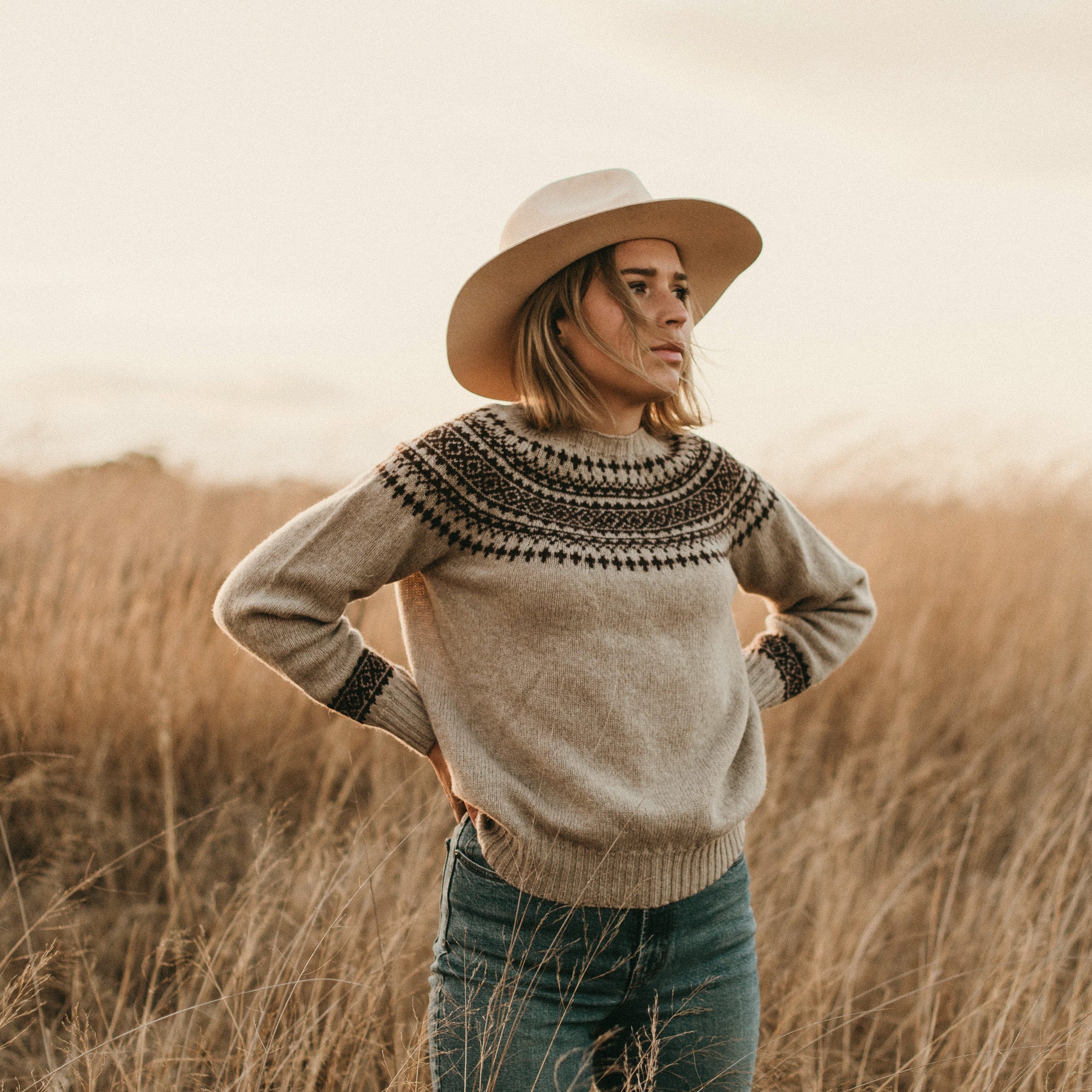 Woman wearing a patterned sweater and hat standing in a field with a neutral background