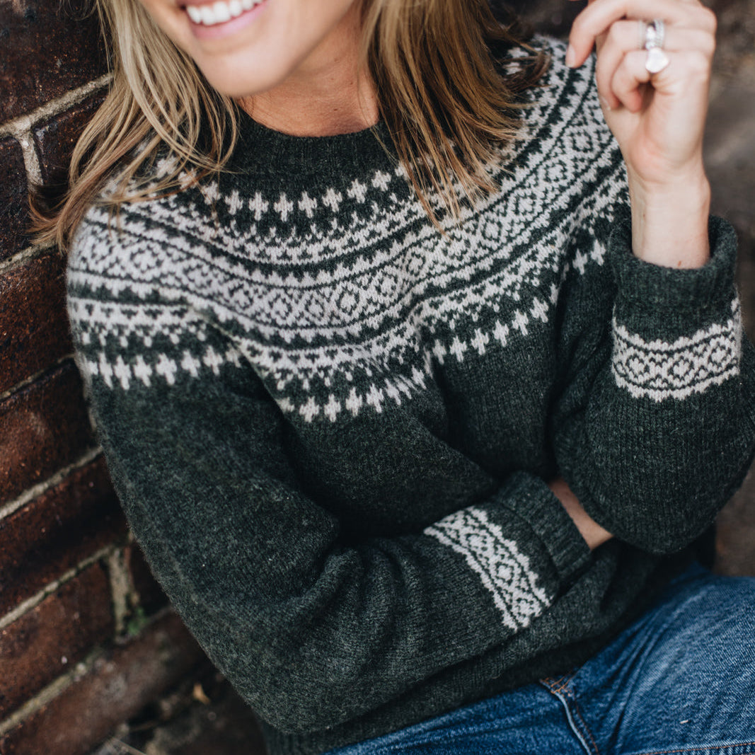 A woman smiling and wearing a dark green fair isle knit sweater with cream-colored patterns on the collar and cuffs.
