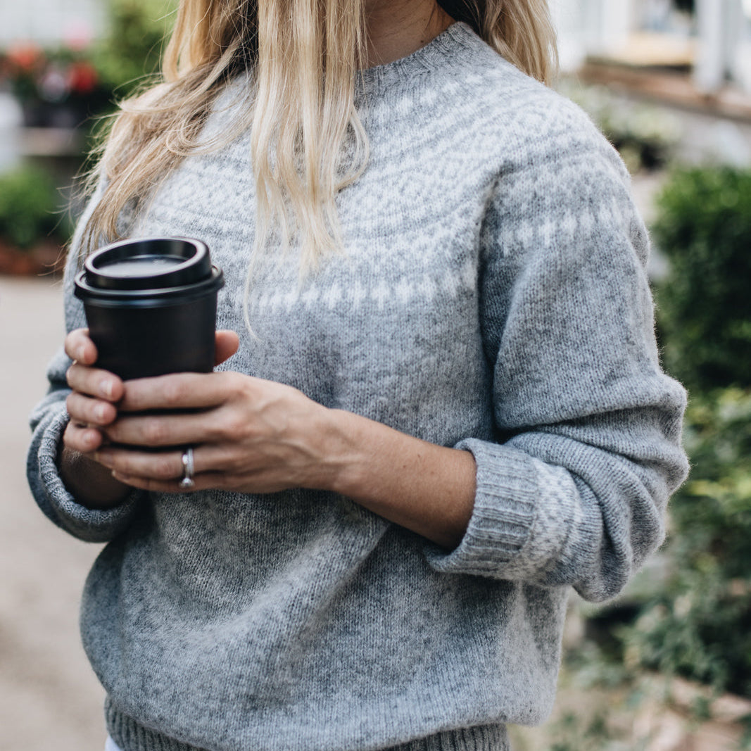 A woman holding a black cup, wearing a silver Fair Isle knit sweater with long sleeves.