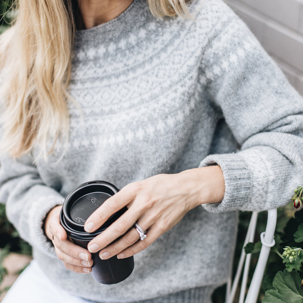 A woman holding a black cup, wearing a silver Fair Isle knit sweater with long sleeves.